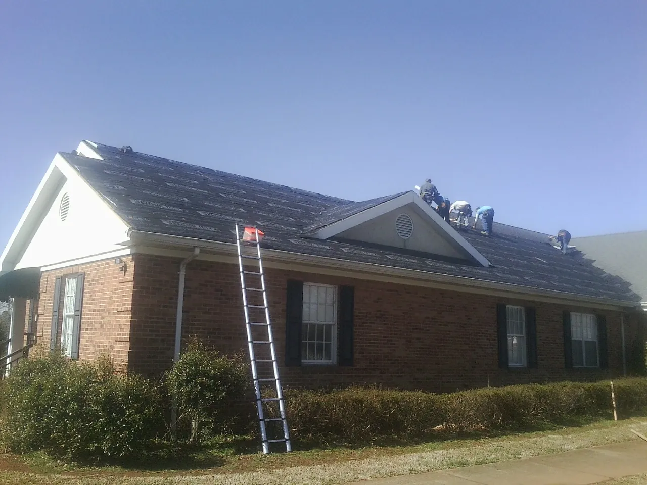 Crew tearing shingles off a brick ranch roof with a ladder set — Emergency Roof Repair work by our crew in Baldwinsville