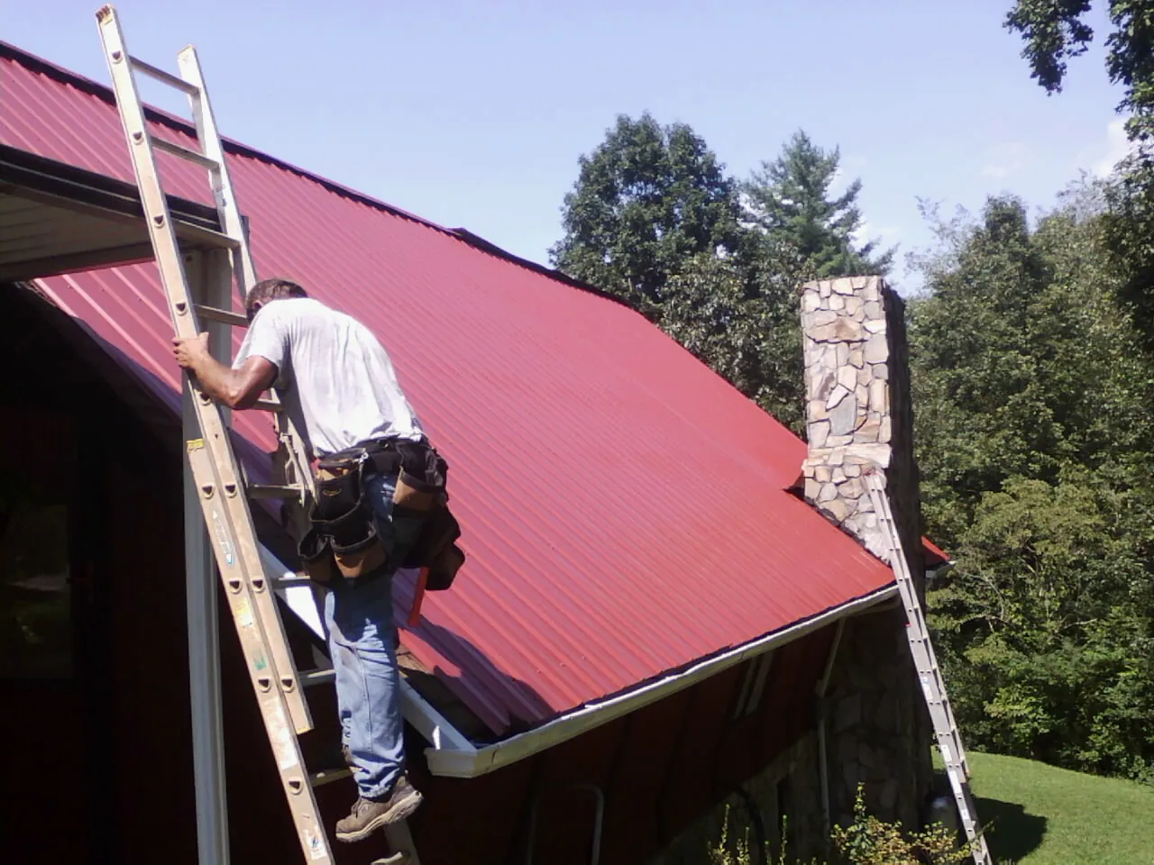 Roofer on a ladder beside a red metal roof and stone chimney — Soffit & Fascia Repair by Roofing Brothers in Baldwinsville